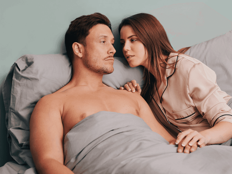 Couple in bed with grey Shreddies anti-flatulence sheets, woman leaning towards man, soft lighting.