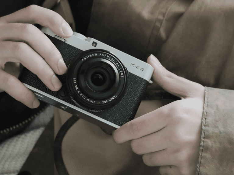 Fujifilm X-E4 camera held by person in brown jacket, highlighting lens and compact design.