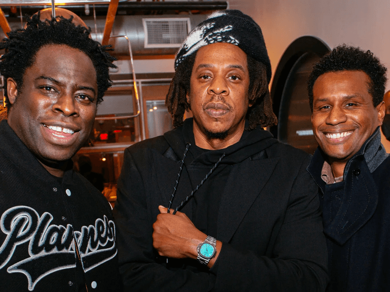 Jay-Z wearing a Patek Philippe Tiffany Nautilus 5711 watch, standing between two smiling men indoors.