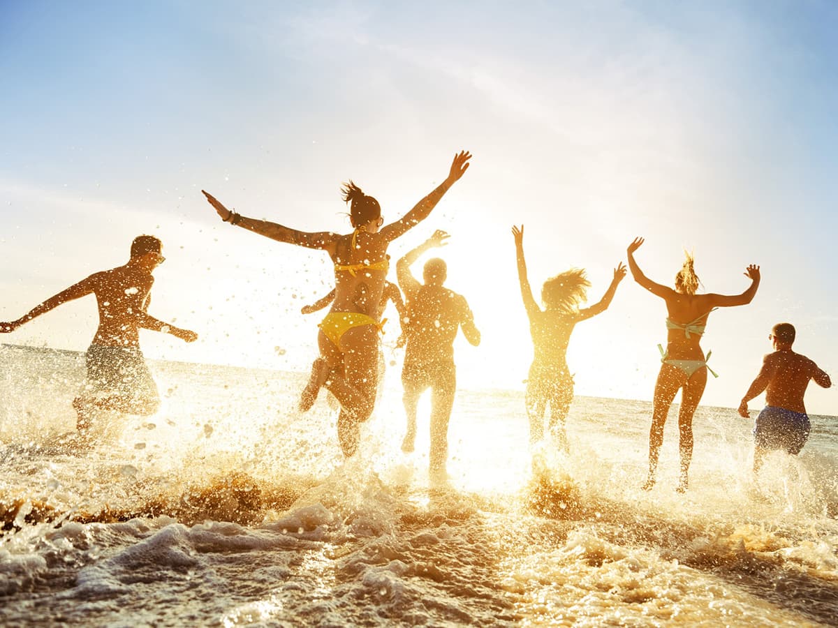 Group of people joyfully splashing in ocean waves at sunset, silhouetted against a bright sky.