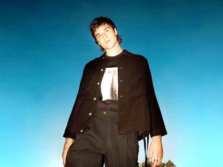 Jacob Elordi wearing a black jacket and graphic t-shirt against a vibrant blue sky.