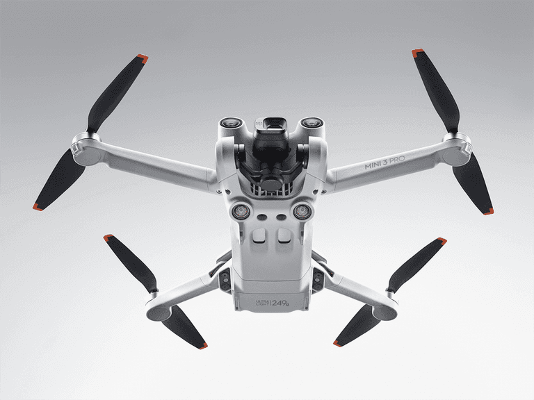 DJI Mini 3 Pro drone in flight with extended propellers against a grey background.