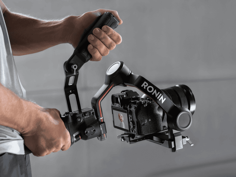 DJI Ronin RS3 gimbal held by person, stabilising a camera with a grey background.