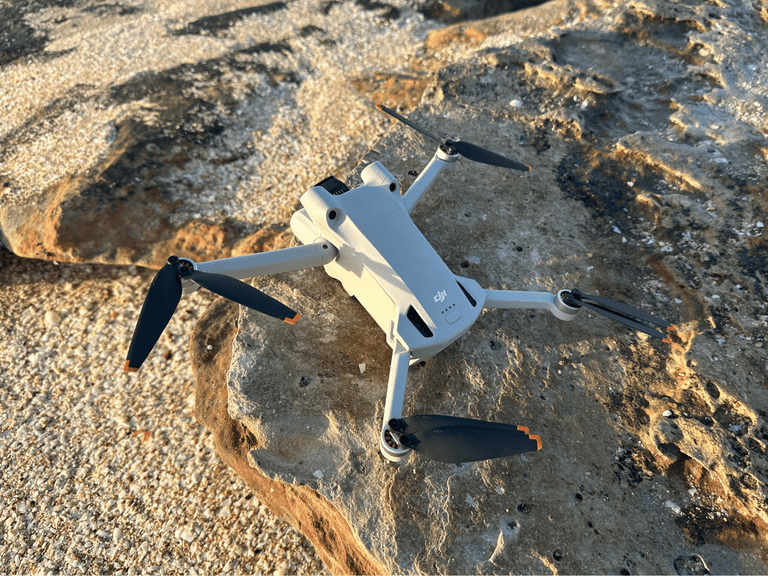 DJI Mini 3 Pro drone on rocky surface with black propellers and sunlight casting shadows.