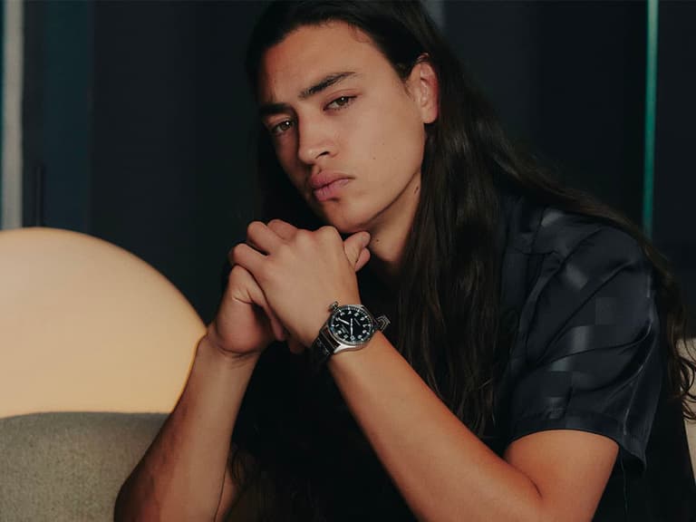 Man with long hair wearing an IWC Big Pilot watch, sitting indoors with hands clasped.