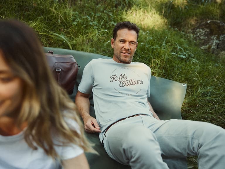 Man in R.M. Williams t-shirt relaxing on grass with leather bag nearby, outdoors in natural light.