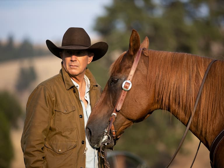 Man in brown jacket and cowboy hat stands beside a chestnut horse in a rural outdoor setting.