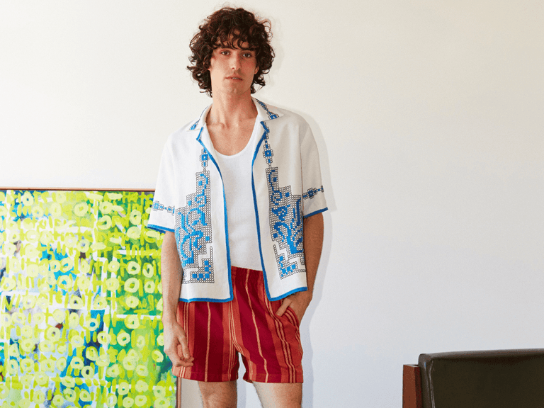Man wearing Hagon Workroom white embroidered shirt and red striped shorts stands indoors near colourful artwork.