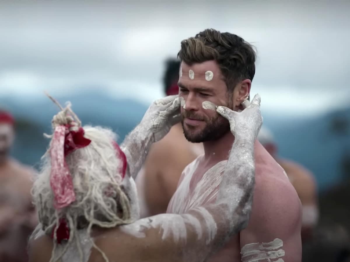 Chris Hemsworth with tribal paint applied to his face and body in an outdoor setting.