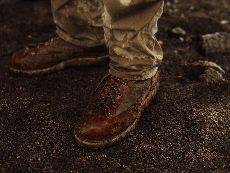 Huckberry x Danner waxed canvas boots on muddy ground, worn with beige trousers.