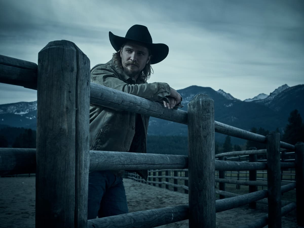 Man in cowboy hat leaning on wooden fence with mountainous landscape in the background under overcast sky.