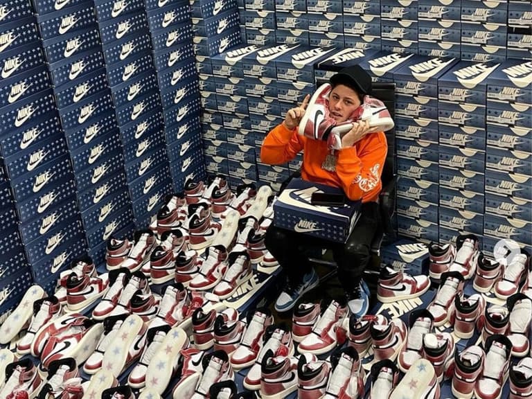 Person in orange hoodie surrounded by stacks of Nike shoe boxes and holding multiple pairs of red and white sneakers.