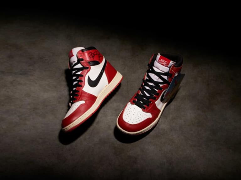 Air Jordan 1 sneakers from 1985 in red, white, and black on a dark surface, with one shoe upright and the other tilted.