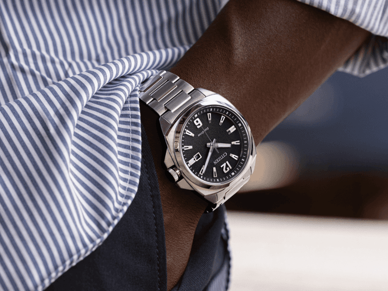 Citizen solar-powered watch with a black dial and silver stainless steel bracelet on a person's wrist, wearing a blue and white striped