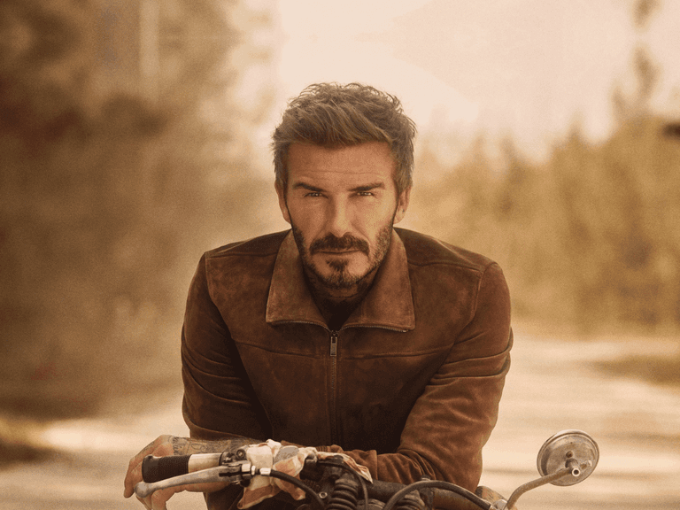 David Beckham wearing a brown suede jacket, sitting on a motorcycle in a sunlit outdoor setting.