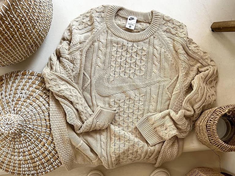 Nike cable knit sweater in beige with intricate patterns, laid on a light surface surrounded by woven baskets.