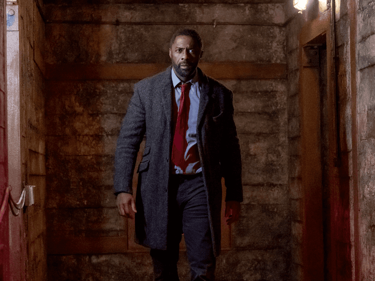 Idris Elba wearing a grey coat and red tie, walking through a dimly lit, rustic hallway in "Luther: The Fallen Sun".