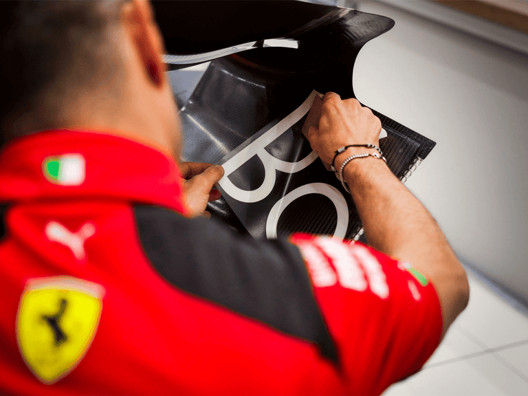 Ferrari team member applies Bang & Olufsen logo to carbon fibre car part, wearing red uniform with Ferrari badge.