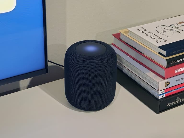 Apple HomePod 2nd Gen in black on a desk beside a stack of books, with soft ambient lighting.