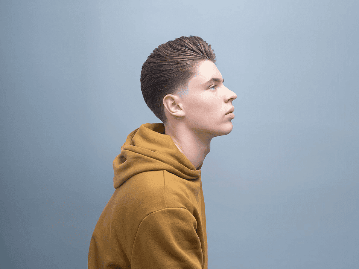 Man with low taper fade hairstyle wearing mustard hoodie against blue background.