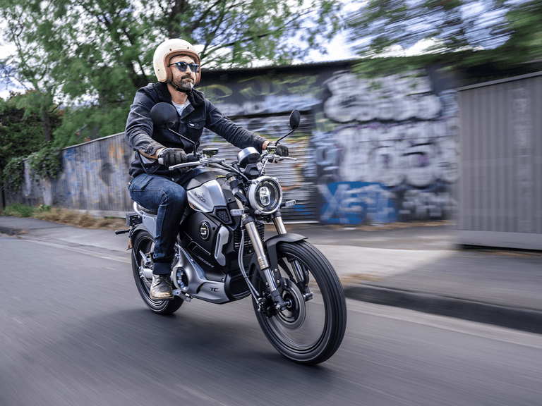 Rider on Super Soco TC-Max wearing helmet and sunglasses, cruising past graffiti-covered wall.