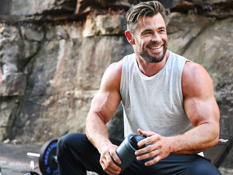 Chris Hemsworth smiling outdoors, wearing a grey tank top and holding a water bottle, with rocky background.