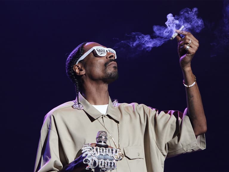 Person wearing sunglasses exhales smoke while holding a lit joint on stage, dressed in a beige shirt.