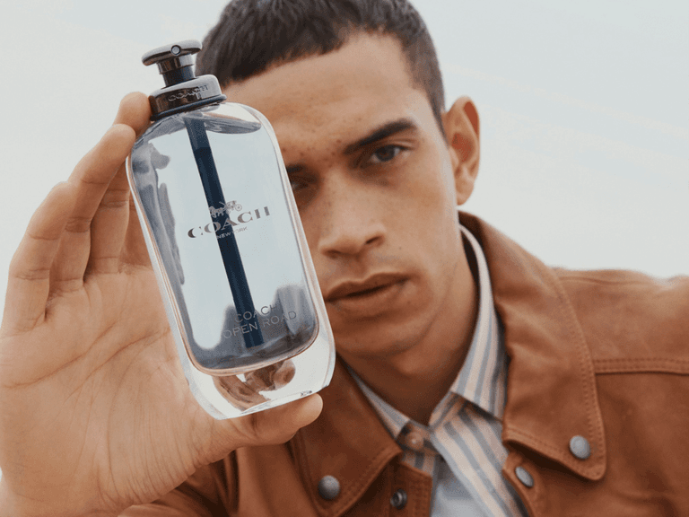 Man holding Coach Open Road fragrance bottle, wearing a brown leather jacket and striped shirt, close-up.