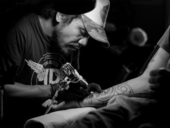 Tattoo artist wearing a cap inks intricate design on a man's forearm in black and white.