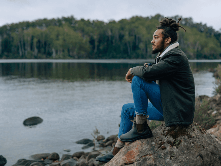 Man sitting on rock by lake wearing Blundstone boots, blue jeans, and a dark jacket, with forested background.