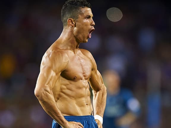 Cristiano Ronaldo shirtless, flexing muscles on a football field with intense expression.