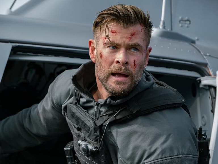 Chris Hemsworth in tactical gear with blood on his face, emerging from a helicopter in an action scene.