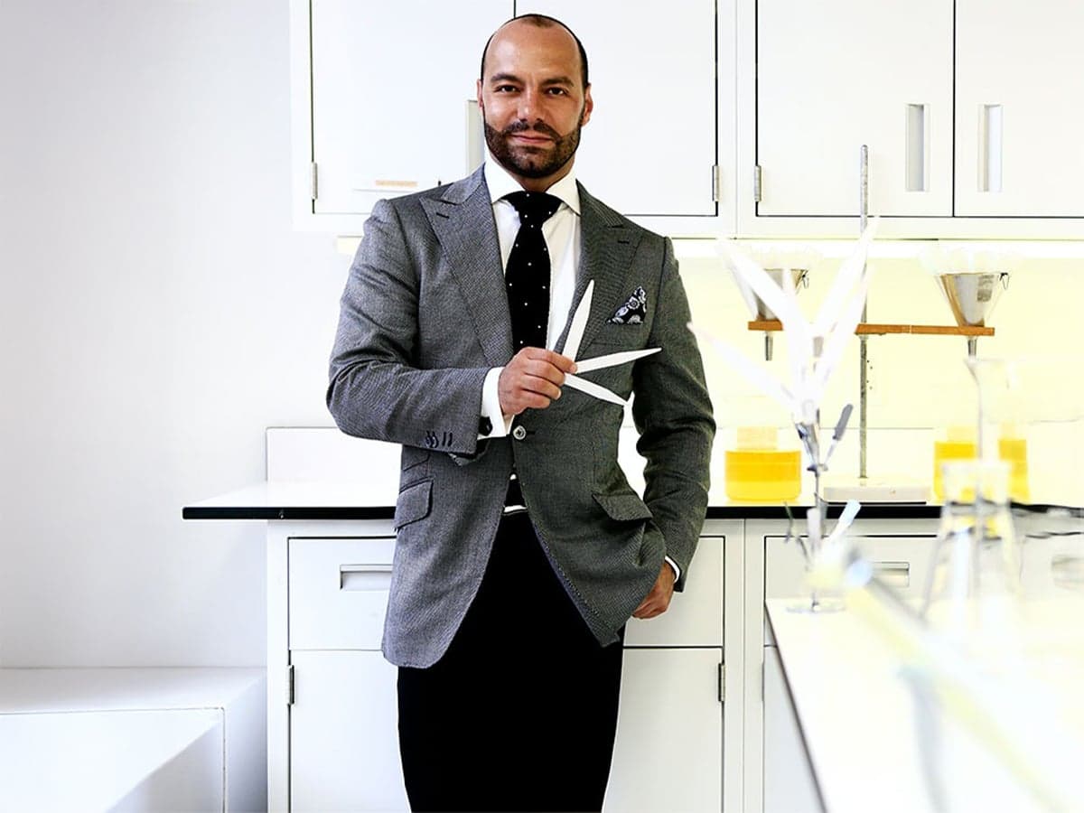 Perfumer in grey suit holding scent strips in a bright, modern laboratory with glass vials and white cabinets.