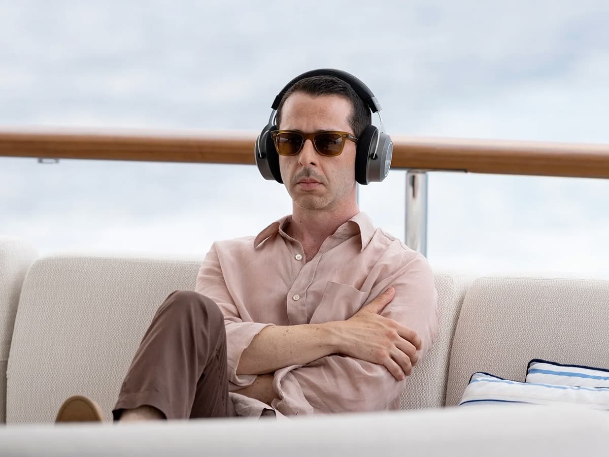 Jeremy Strong as Kendall Roy wearing sunglasses and headphones, seated on a yacht in a light pink shirt and brown pants.