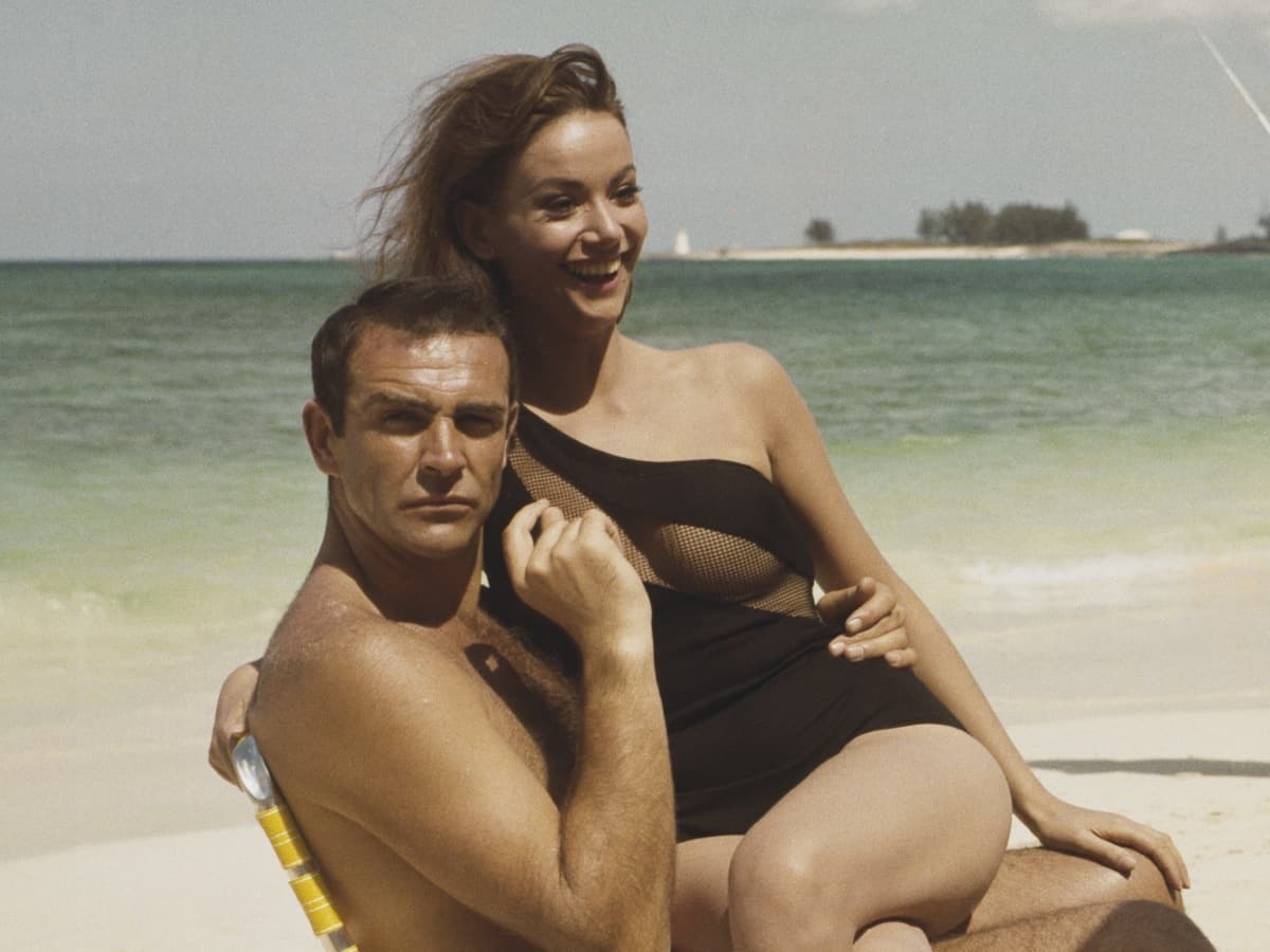 Claudine Auger in black swimsuit sits on Sean Connery's lap on a beach, both smiling, with ocean in background.