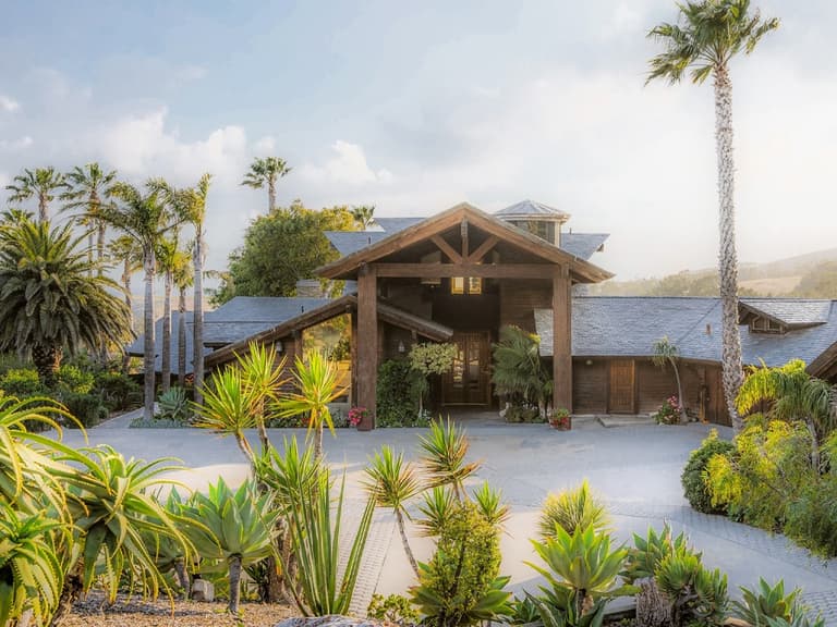James Cameron's California ranch entrance with lush greenery, palm trees, and a rustic wooden facade under a clear sky.