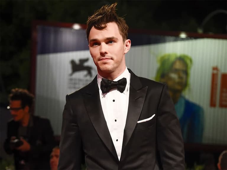 Man in black tuxedo with bow tie and unique hairstyle at an event, blurred background.
