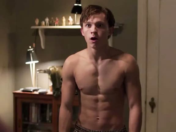 Tom Holland standing shirtless in a dimly lit room, showing defined abs and a surprised expression.