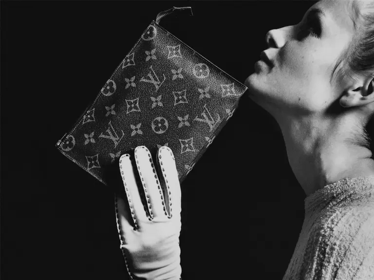 Woman holding a Louis Vuitton purse with a gloved hand against a dark background.