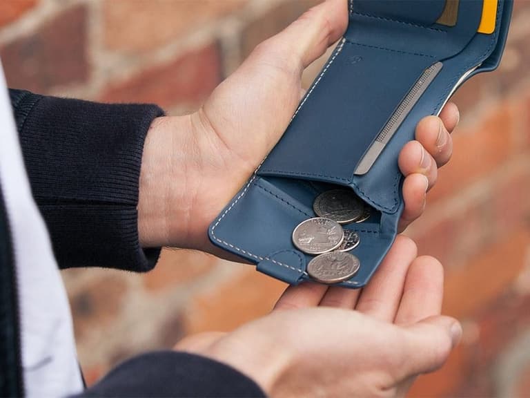 Hands holding a blue bi-fold wallet with coins and a card against a brick wall background.