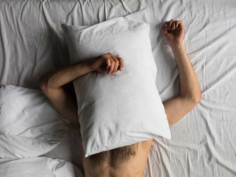 Man lying on white bed with pillow covering his face, arms resting beside his head.