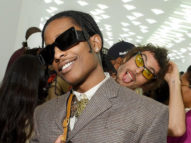A man in a patterned brown suit and oversized sunglasses smiles at an indoor event, with another person in yellow glasses behind him.