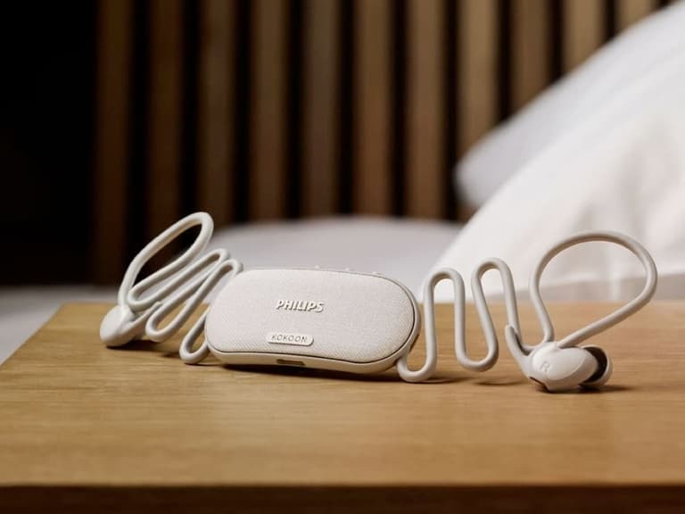 Philips sleep earbuds in white on a wooden bedside table, with a sleek charging case and coiled cables.
