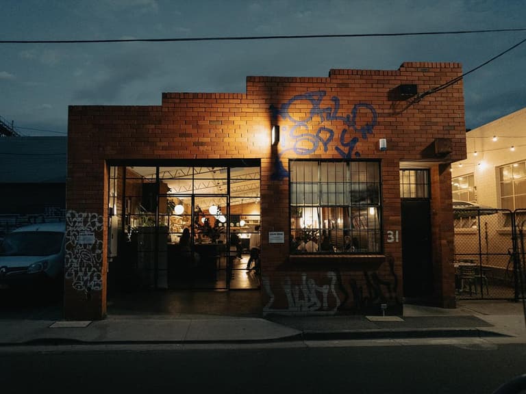 Brick building with graffiti, warm interior lighting visible through open doors, evening setting.