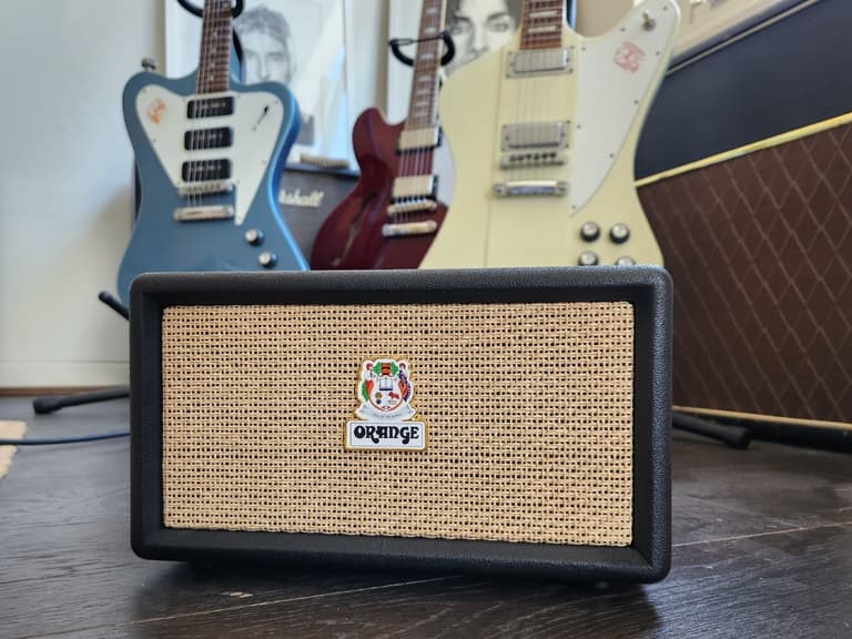 Orange Box Bluetooth speaker with woven front grille on dark wood floor, guitars and amplifier in background.