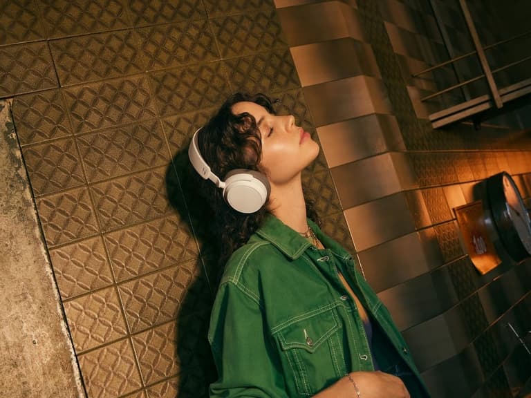 Person wearing Sennheiser Accentum wireless headphones in white, leaning against a patterned wall with warm lighting.