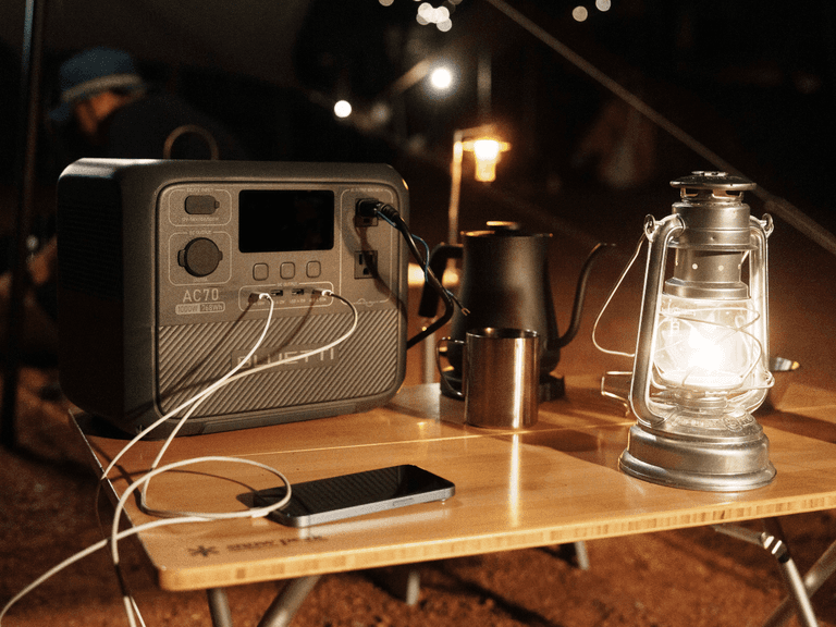 Bluetti AC70 portable generator on a wooden table with a lantern, smartphone, and cables at night.