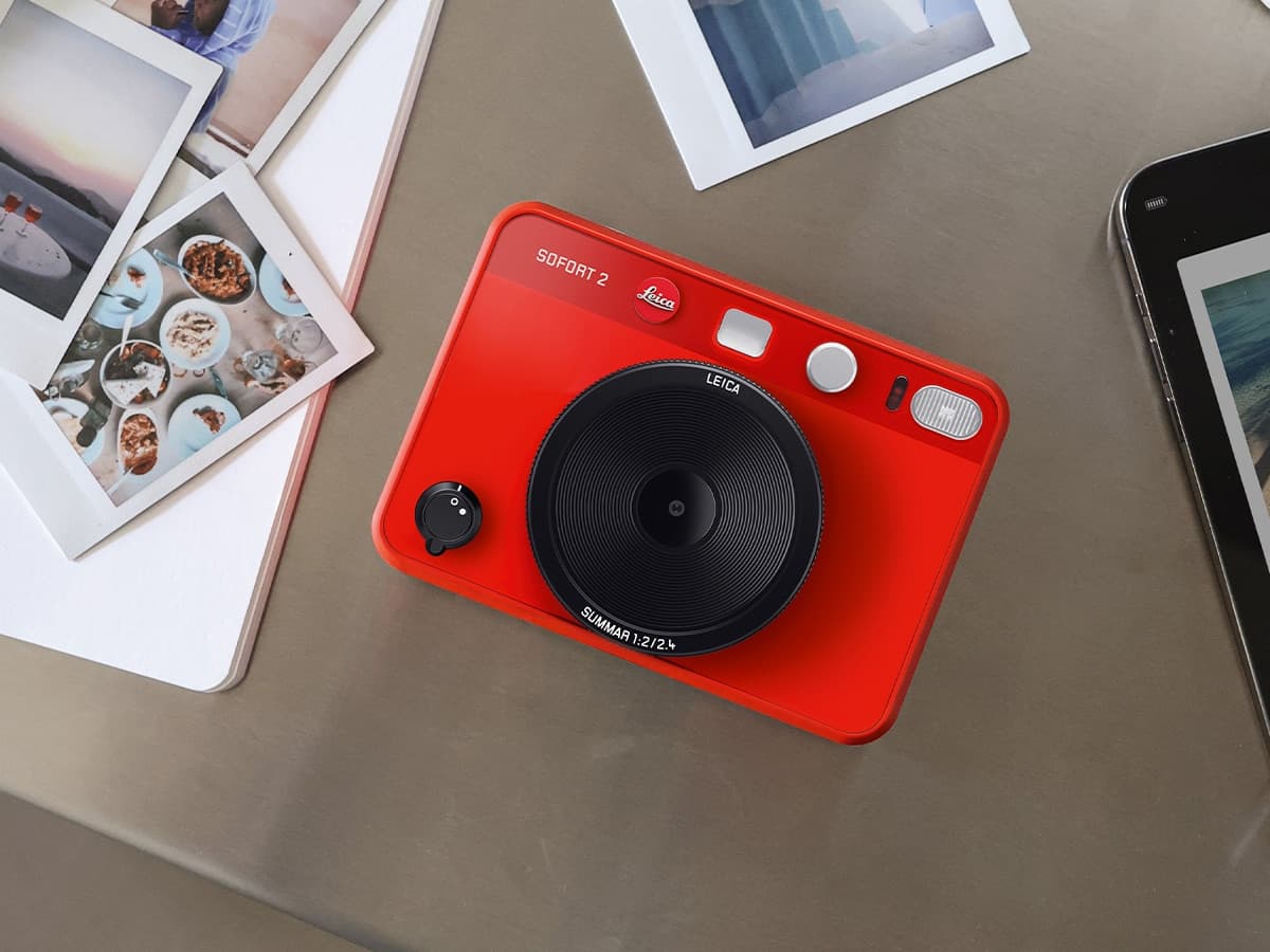 Leica Sofort 2 camera in vibrant red on a table with scattered photos and a smartphone nearby.