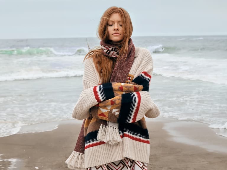 Woman wearing Tommy Hilfiger x Pendleton knitwear stands on a beach, wrapped in a patterned scarf, with waves in the background.