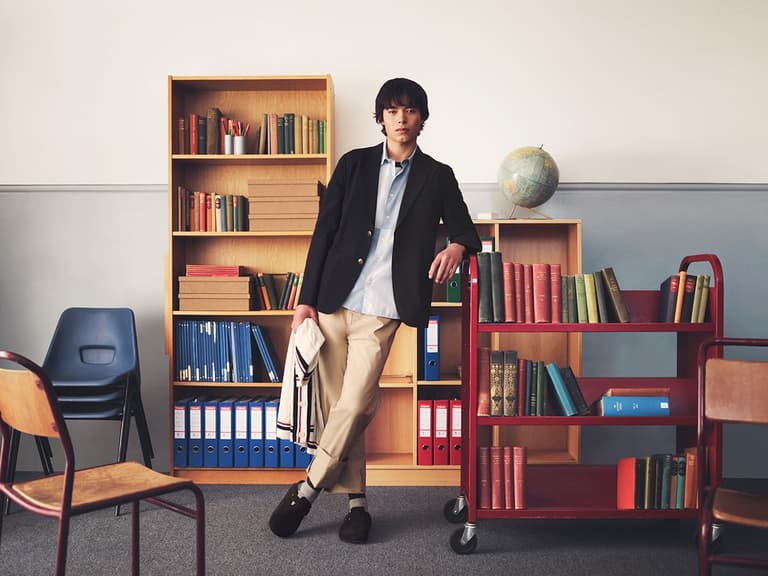 Man in Uniqlo x JW Anderson outfit leaning on bookshelf in classroom setting, wearing a navy blazer and beige trousers.